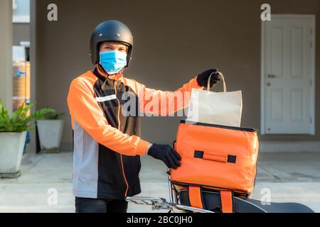 Lieferung Asian man tragen Schutzmaske in orangefarbener Uniform und sind bereit, Liefer-Food-Tüte vor den Kundenhouten mit Etui auf Roller zu schicken, z. Stockfoto