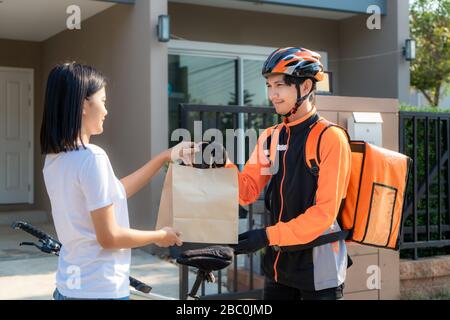 Asian man Courier auf dem Fahrrad, der Lebensmittel in einem orangefarbenen, einheitlichen Lächeln liefert und Lebensmittelbeutel im Vorderhaus hält und Asian Woman eine Lieferung von Kisten annimmt Stockfoto