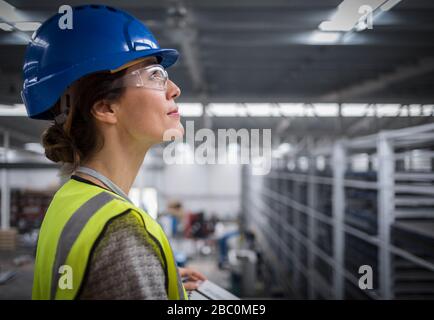 Lächelnde, selbstbewusste weibliche Führungskraft, die in der Fabrik nach einem Hut Ausschau halten muss Stockfoto