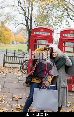 Seniorinnen Freunden sich mit Einkaufstaschen an, die selfie im Herbstpark vor roten Telefonzellen einnehmen Stockfoto