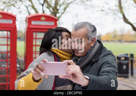 Senioren-Paar küsst und nimmt selfie in Park vor roten Telefonzellen Stockfoto