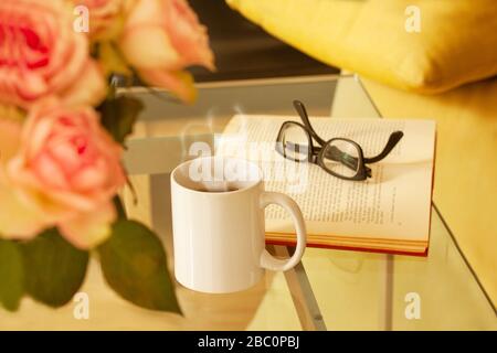 Buch, Brille, Rosen und ein Becher mit Tee auf modernem Glastisch. Wohnzimmer Inneneinrichtung Inneneinrichtung Konzept. Stockfoto