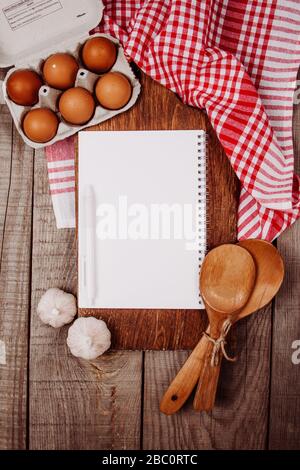 Holzschneidebrett auf Holzgrund mit Knoblauch, Pfannen, Eiern und einem leeren Notizbuch Stockfoto