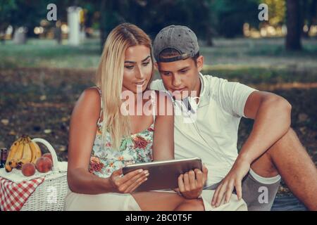 Schuss eines glücklichen jungen Paares, das beim Picknick im Park etwas auf einem Tablet beobachtet. Stockfoto