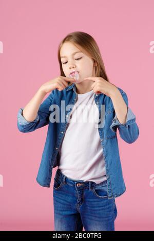 Kleines Mädchen mit Sonnenbrille, das rosafarbenen Kaugummi vor einem hellvioletten Hintergrund aufbläst. Stockfoto