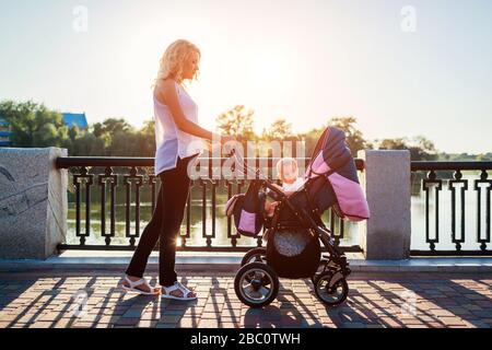 Muttertag. Junge Mutter, die im Frühlingspark mit Kleinkind in der Babykutsche spazieren geht. Frau verbringt Zeit mit Kind im Freien Stockfoto