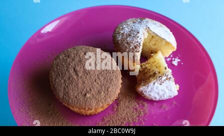 Zwei mit Schokolade und Puderzucker bestreute Quark auf einem rosafarbenen Teller, auf blauem Grund. Dessert, ein kleiner Cupcake. Weiße, gebackene Plätzchen mit einem Stockfoto