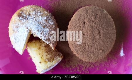 Zwei mit Schokolade und Puderzucker bestreute Quark auf einem rosafarbenen Teller, auf blauem Grund. Dessert, ein kleiner Cupcake. Weiße, gebackene Plätzchen mit einem Stockfoto