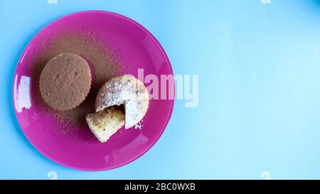 Zwei mit Schokolade und Puderzucker bestreute Quark auf einem rosafarbenen Teller, auf blauem Grund. Dessert, ein kleiner Cupcake. Weiße, gebackene Plätzchen mit einem Stockfoto