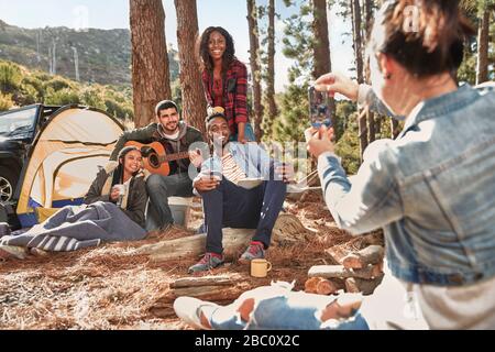 Junge Frau mit Kameratelefon fotografiert Freunde, die auf dem Zeltplatz im Wald hängen Stockfoto