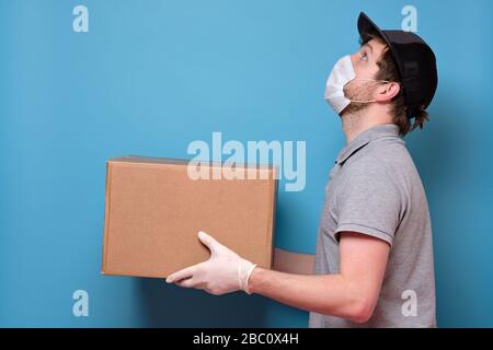 Kaukasischer junger Mann in medizinischer Maske, der ein Paket hält, das er dem Kunden gibt. Kein ansteckendes Lieferkonzept. Studio an blauer Wand gedreht. Stockfoto
