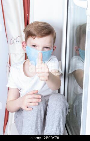 Selbstisolierungs- und Quarantänekonzept. Ein siebenjähriger Junge in einer medizinischen Maske sitzt am Fenster und hält den Daumen hoch Stockfoto