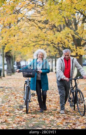 Älteres Paar, das im Herbstpark Fahrräder zwischen Bäumen und Blättern spazieren geht Stockfoto