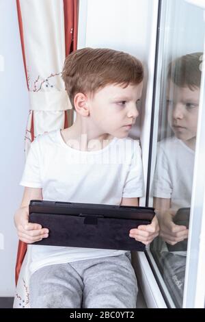 Fernunterricht im Selbstisolierungsmodus und Quarantänekonzept. Ein siebenjähriger Junge sitzt am Fenster und hält eine Tablette in den Händen. Online Stockfoto