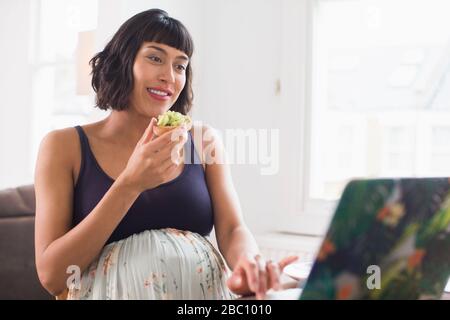 Glückliche schwangere Frau, die Avocado Toast auf dem Laptop isst Stockfoto