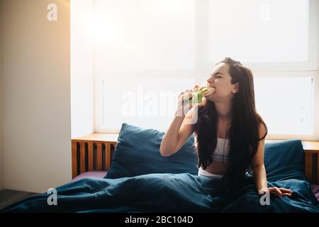 Junge schöne Frau im Morgenbett zu Hause. Sandwich mit grünem Gemüse und leckeren Broten essen und beißen. Frühstück zu Hause. Stockfoto