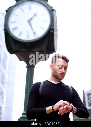 Besorgter Geschäftsmann, der unter der Uhr der Stadt in einer Innenstadt steht und seine Armbanduhr, das Konzept eines Zeitmanagements und der Hurrying betrachtet. Stockfoto