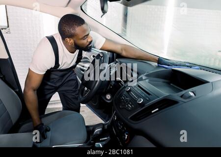 Automatische Detaillierung und Valeting-Konzept. Gutaussehender afroamerikanischer Mitarbeiter für den Autoservice, mit weißem T-Shirt und grauen Overalls, Reinigungswagen im Innenraum Stockfoto