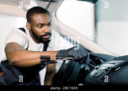 Junger, gutaussehender, konzentrierter afroamerikanischer Mann mit Reinigungsbürste und Staubentfernung vom Lüftungsgitter der Autoklimaanlage. Autodetails oder Stockfoto