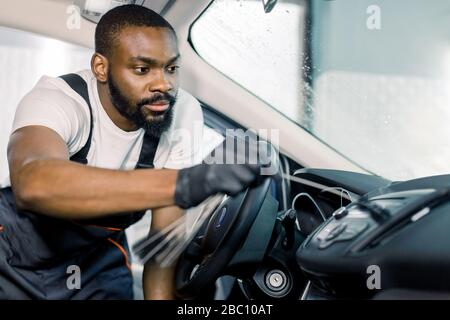 Afrikanischer Mann, professioneller Autodienstmitarbeiter mit schwarzen Schutzhandschuhen, mit speziellen Bürsten im Innenraum reinigen. Automatische Detaillierung und Valeting Stockfoto