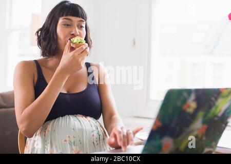 Schwangere Frau, die Avocado Toast auf dem Laptop isst Stockfoto