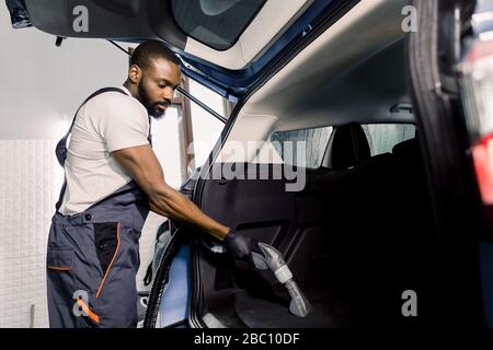 Afrikanischer männlicher Arbeiter, der weißes T-Shirt und graue Overalls trägt, im Innenraum des Autos saugt, Kofferraum mit Nassstaubsauger, professionelle Absaugung Stockfoto