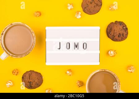 Kürzel Wort JOMO auf einer dekorativen Tafel auf hellgelbem Hintergrund. Das Konzept der Entspannung durch Informationen und Geräte. Draufsicht, flacher Lay. Stockfoto