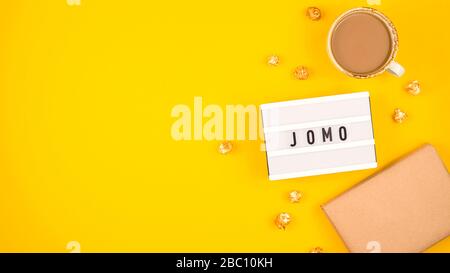 Kürzel Wort JOMO auf einer dekorativen Tafel auf hellgelbem Hintergrund. Das Konzept der Entspannung durch Informationen und Geräte. Draufsicht, flaches Layout, Kopierbereich. Stockfoto
