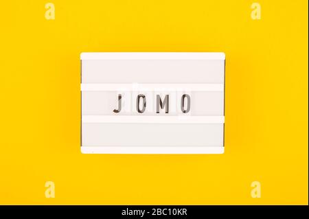Kürzel Wort JOMO auf einer dekorativen Tafel auf hellgelbem Hintergrund. Das Konzept der Entspannung durch Informationen und Geräte. Draufsicht, flacher Lay. Stockfoto