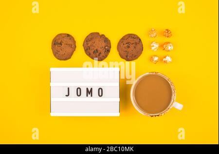 Kürzel Wort JOMO auf einer dekorativen Tafel auf hellgelbem Hintergrund. Das Konzept der Entspannung durch Informationen und Geräte. Draufsicht, flacher Lay. Stockfoto