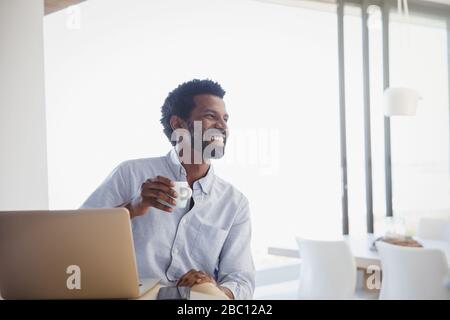 Lächelnder, begeisterter Mann, der Kaffee trinkt und zu Hause am Laptop arbeitet Stockfoto