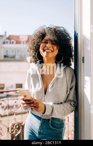 Porträt der glücklichen jungen Frau mit lockigen Haaren hält Handy auf dem Balkon Stockfoto
