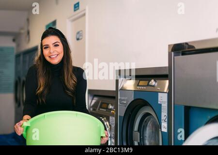 Porträt einer lächelnden jungen Frau, die die Wäsche in einem Waschsalon hält Korb Stockfoto