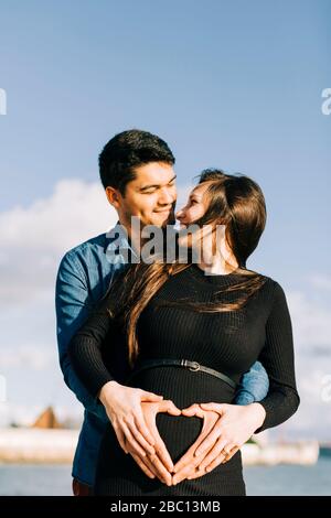 Liebevolle werdende Eltern, die mit ihren Händen am Babybauch ein Herz Formen Stockfoto
