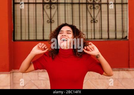 Nahaufnahme des Porträts der glücklichen Brasilianerin mit rotem Kleid, lockerem lockerem lockerem Haar und urbanem Hintergrund in Granada, Spanien Stockfoto