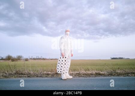 Mann mit Schutzanzug und Maske auf Landstraße mit Toilettenrollen stehend Stockfoto