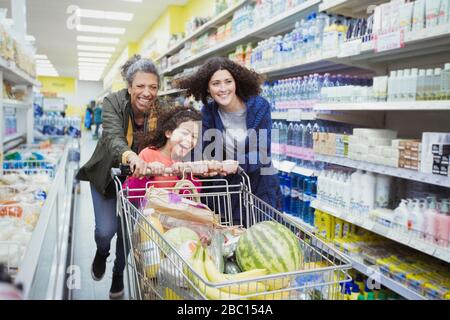 Verspielte Mehrgenerationsfrauen schieben Einkaufswagen in Supermarkt-Gang Stockfoto