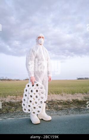 Mann mit Schutzanzug und Maske auf Landstraße mit Toilettenrollen stehend Stockfoto