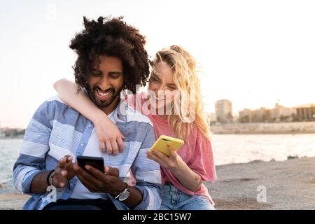 Glückliches junges Paar, das bei Sonnenuntergang mit dem Smartphone auf der Kaimauer sitzt Stockfoto