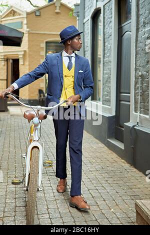 Stilvoller junger Geschäftsmann mit Fahrrad in altmodischem Anzug, der eine Gasse entlang läuft Stockfoto