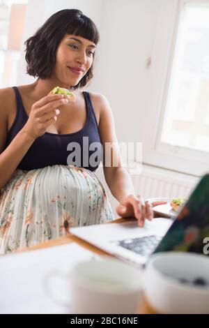 Schwangere Frau, die am Laptop isst Stockfoto