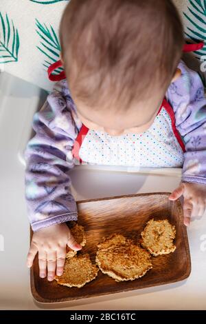 Baby Mädchen sitzt im Hochstuhl essen hausgemachte Haferflocken Cookies mit Händen, Draufsicht Stockfoto