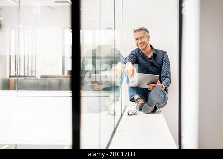 Zwangloser Geschäftsmann sitzt auf Fensterbank im Bürogebäude, mit Laptp Stockfoto