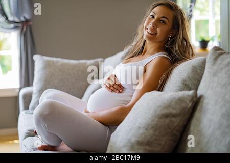 Fröhliche, schöne schwangere Frau, die auf dem Sofa sitzt und ihren Bauch berührt Stockfoto
