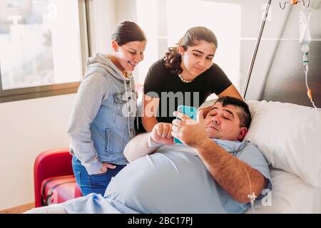 Familie, die Mann mit Handy im Krankenhausbett liegt Stockfoto