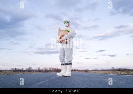 Mann, der Schutzanzug und Maske trägt, hält Lebensmitteltasche auf einer Landstraße Stockfoto