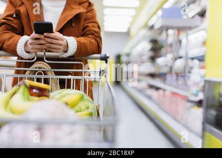 Frau mit Smartphone drückt Einkaufswagen im Supermarkt Stockfoto