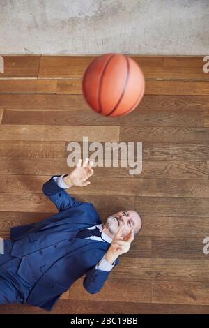 Älterer Geschäftsmann, der auf Holzboden liegt und mit Basketball spielt Stockfoto