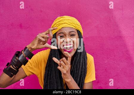 Porträt einer Frau mit langen Dreadlocks, die vorgibt, mit den Händen vor einer rosa Wand ein Foto zu machen Stockfoto