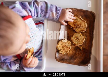 Baby Mädchen sitzt im Hochstuhl essen hausgemachte Haferflocken Cookies mit Händen, Draufsicht Stockfoto